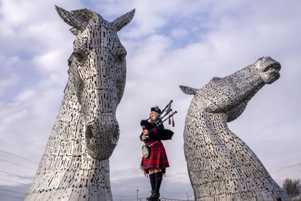 A bagpiper stands between the two metal horses heads of The Kelpies sculpture in Falkirk, playing the bagpipes