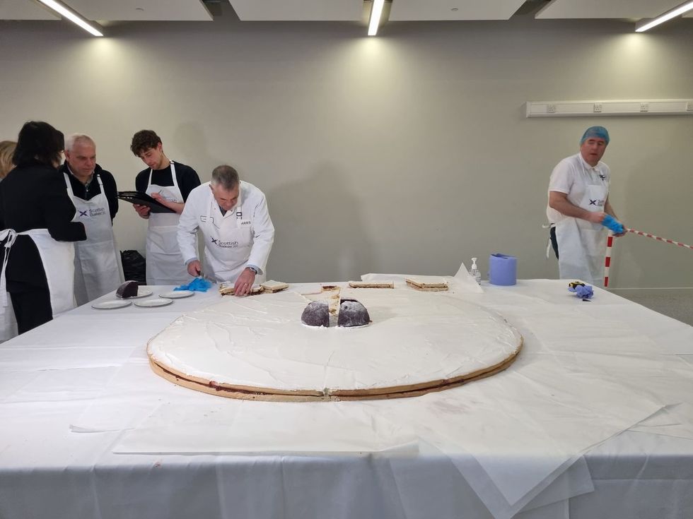 A baker cuts into a giant Empire biscuit, while judges stand nearby
