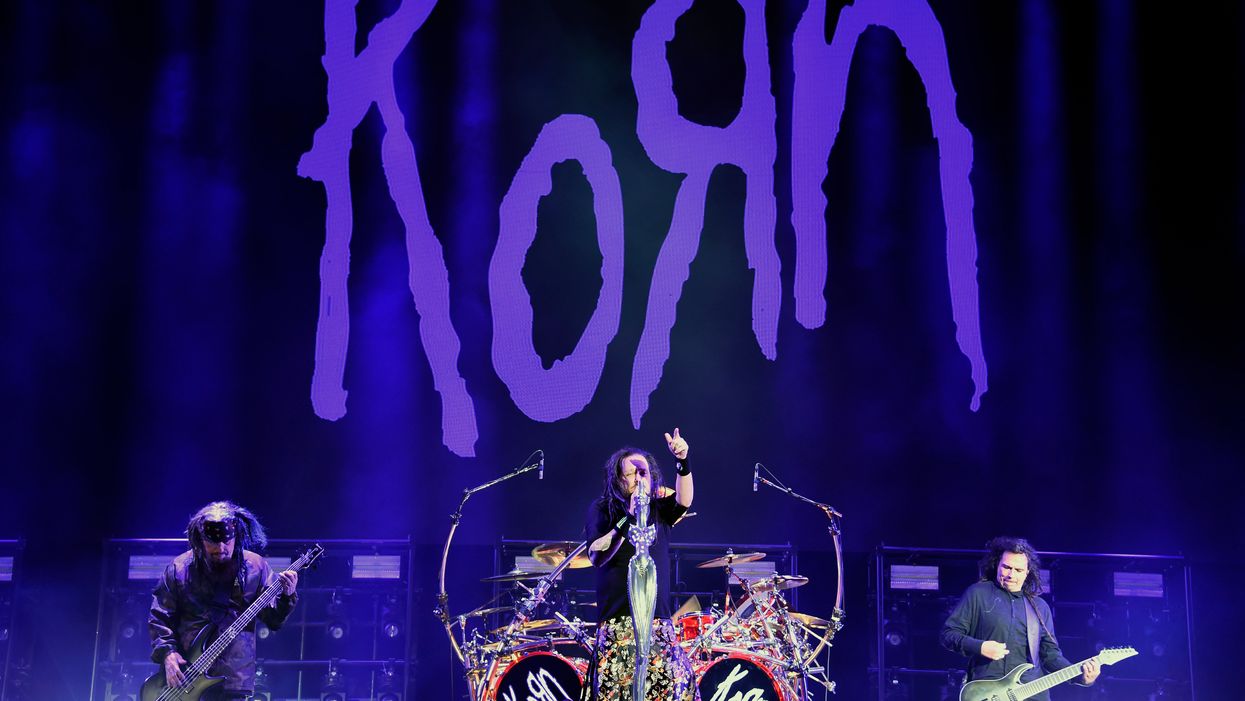 A band on a stage. There’s two guitarists and a drummer in the middle. A banner behind them reads ‘Korn’, with the R flipped horizontally.
