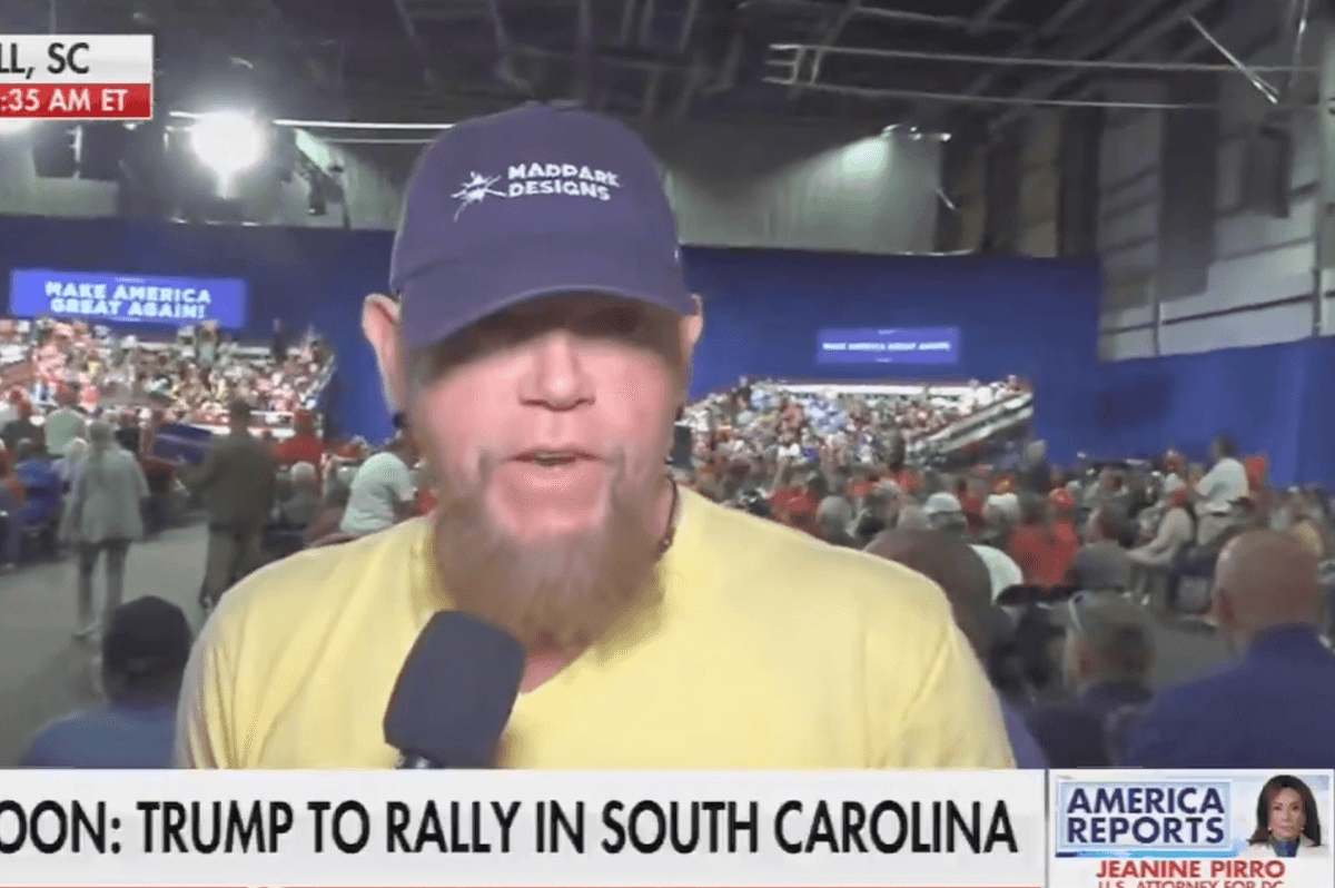 A bearded white man with a blue cap and yellow T-shirt speaks directly to the camera as a journalist holds a microphone to his right.