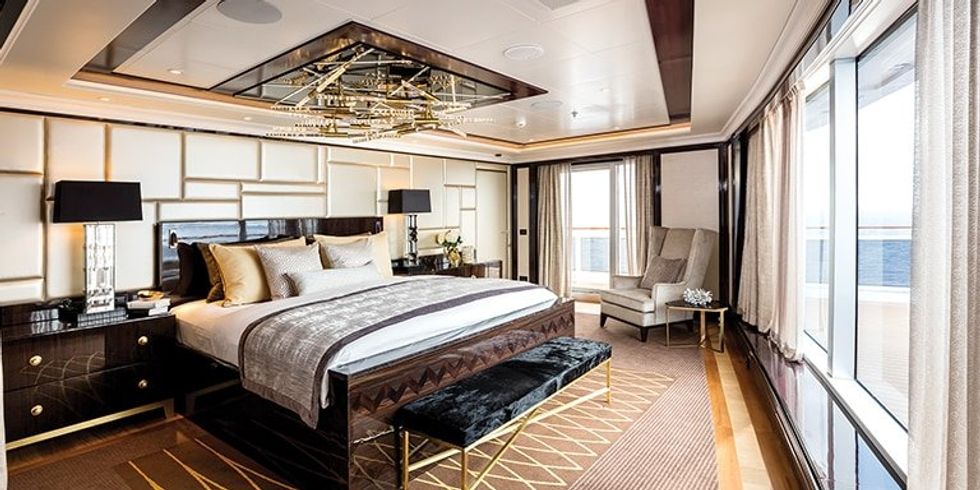 A bedroom in the Regent Suite on board the Seven Seas Splendor. It has a large brown bed and a foot stool, alongside a chair in the corner.