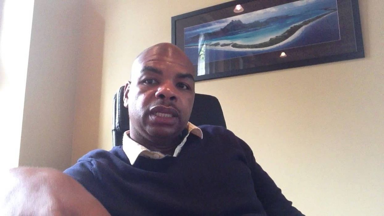 A Black man wearing a blue jumper and white collared shirt sits in his office, talking directly to the camera.