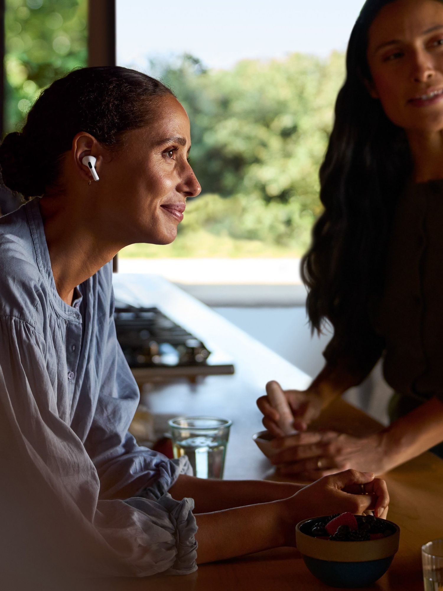 A Black woman with black hair tied back and white AirPods in her ears, smiles at someone off-camera as she rests on a wooden kitchen counter. A Black woman on the right, on the opposite side of the table and with longer black hair, is smiling at someone to her left. A bearded Black man can be seen, out of focus, behind the woman on the left.