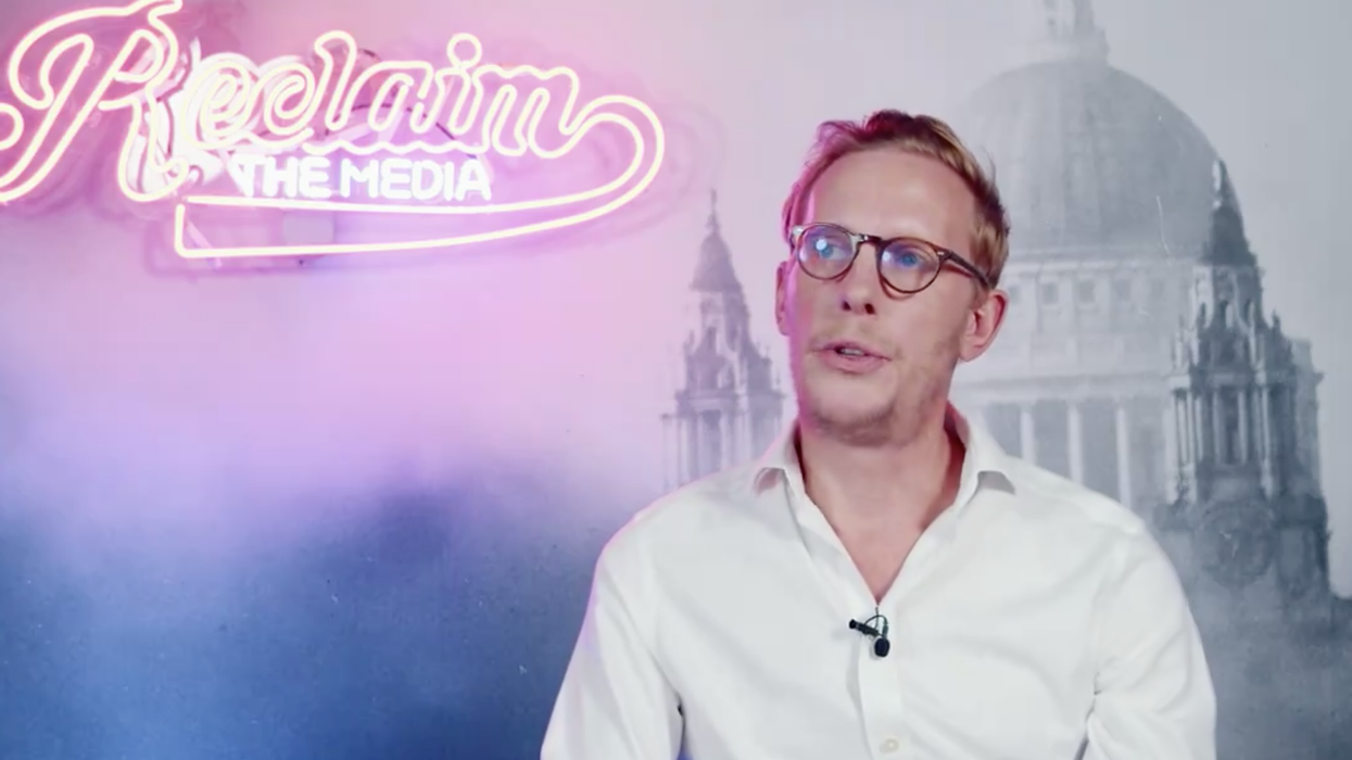 A blond-haired man in glasses in front of a neon sign
