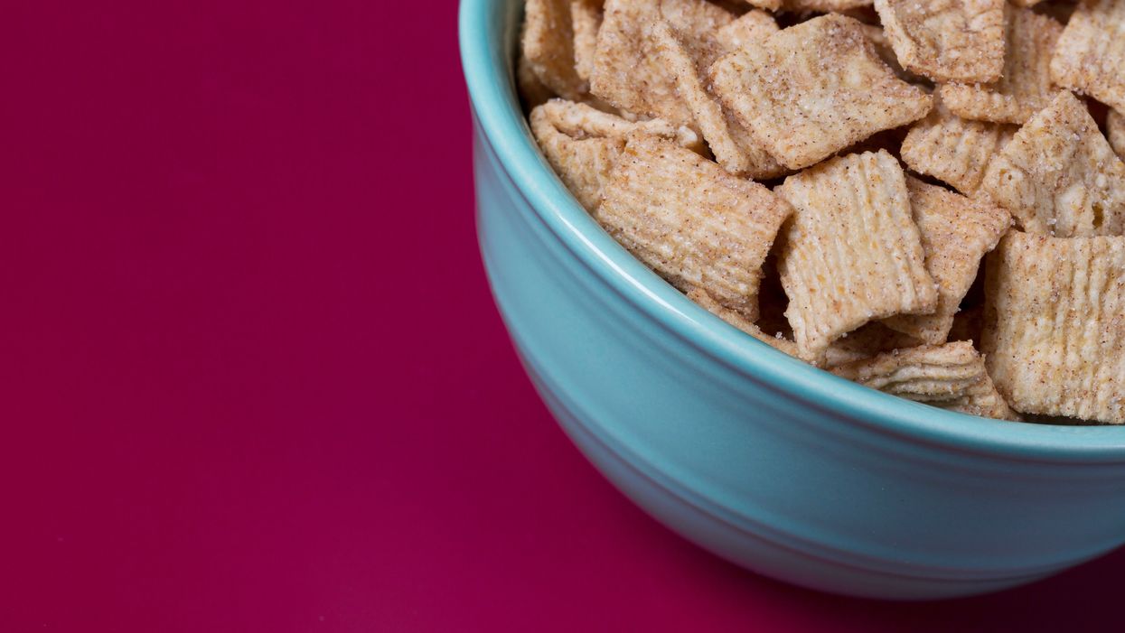 A blue bowl containing cinnamon-coated square breakfast cereal, against a magenta background.