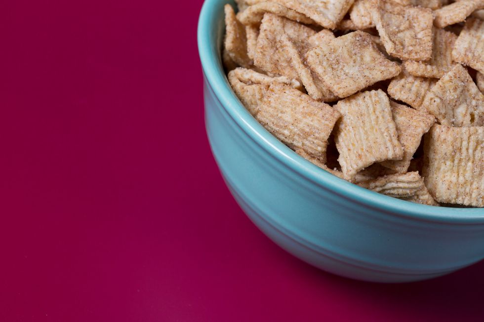 A blue bowl containing cinnamon-coated square breakfast cereal, against a magenta background.