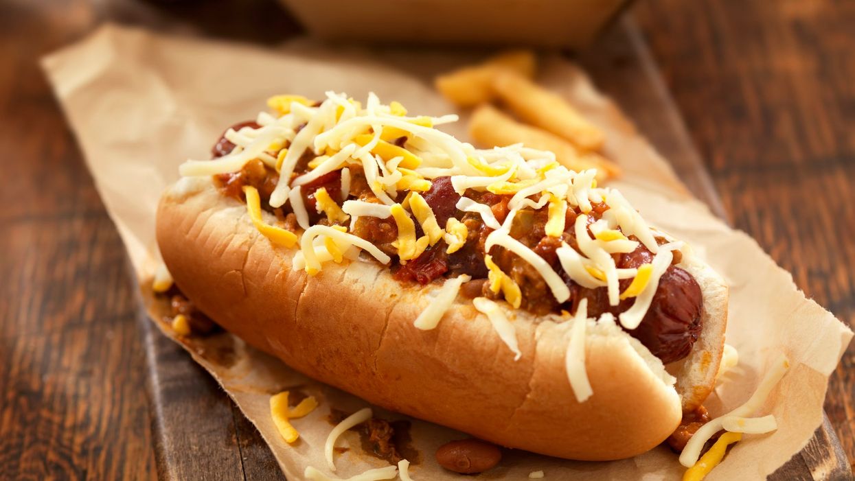 A chilli cheese hot dog resting on a wooden table, with a basket of fries in the background behind it.