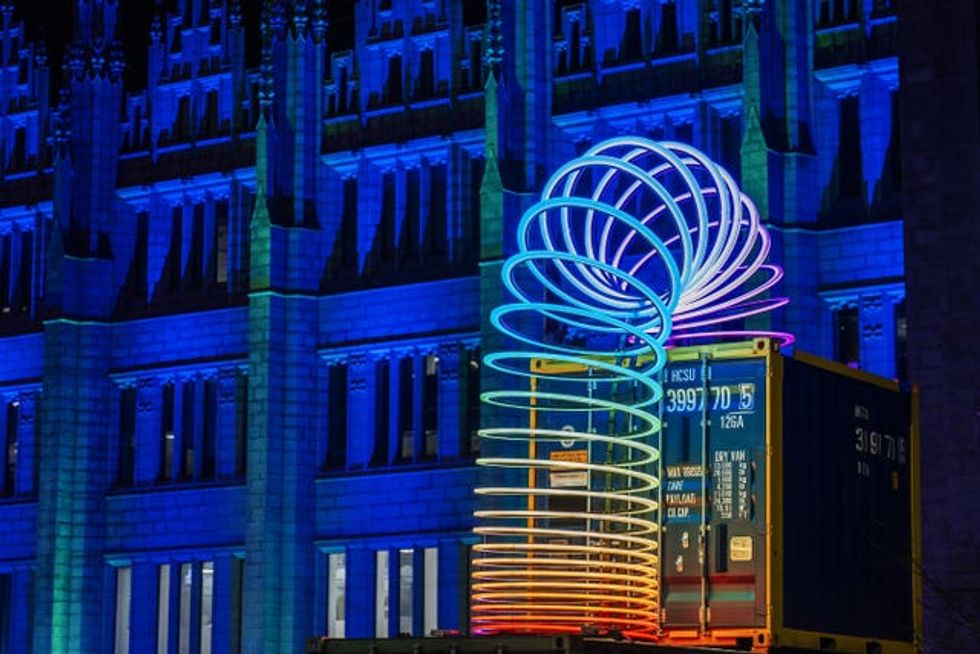 A giant illuminated slinky, on top of a contained in front of a building lit up in blue