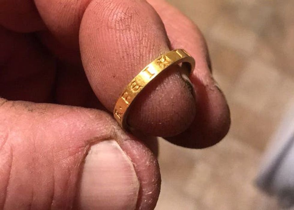 A gold ring with letters inscribed on it, being held on the fingertip of a hand