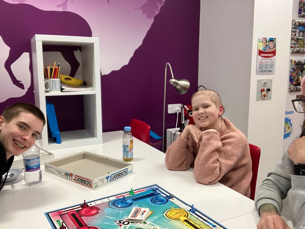 A group of three young people sitting around a table with a board game in the centre. The background features a purple wall