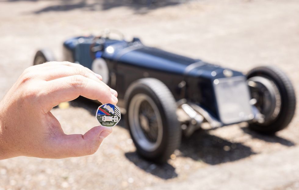 A hand holding a new 50p coin, in front of a navy Delage car