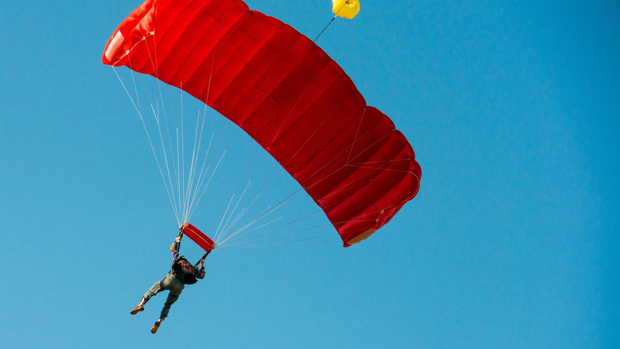 A low angle view of a skydiver in the sky. Their red parachute is deployed and they are preparing to land.