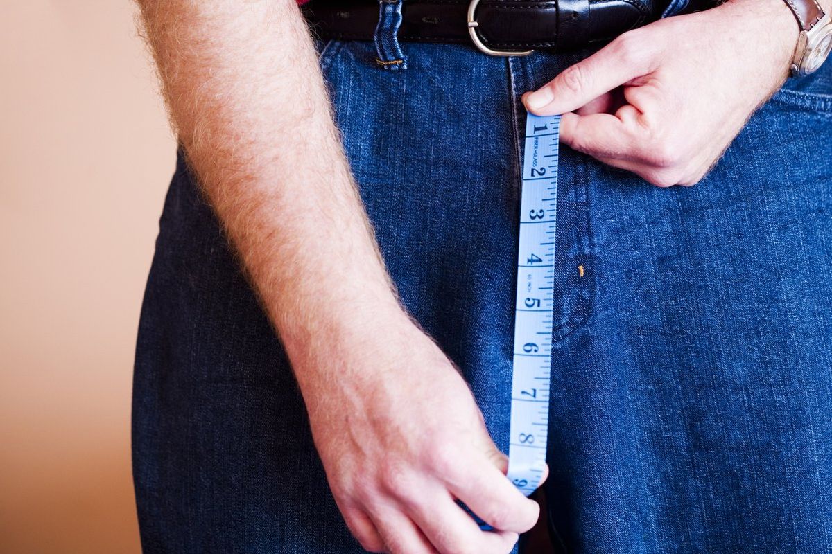 A man holds a tape measure in the region of his groin, implying that he's measuring his penis