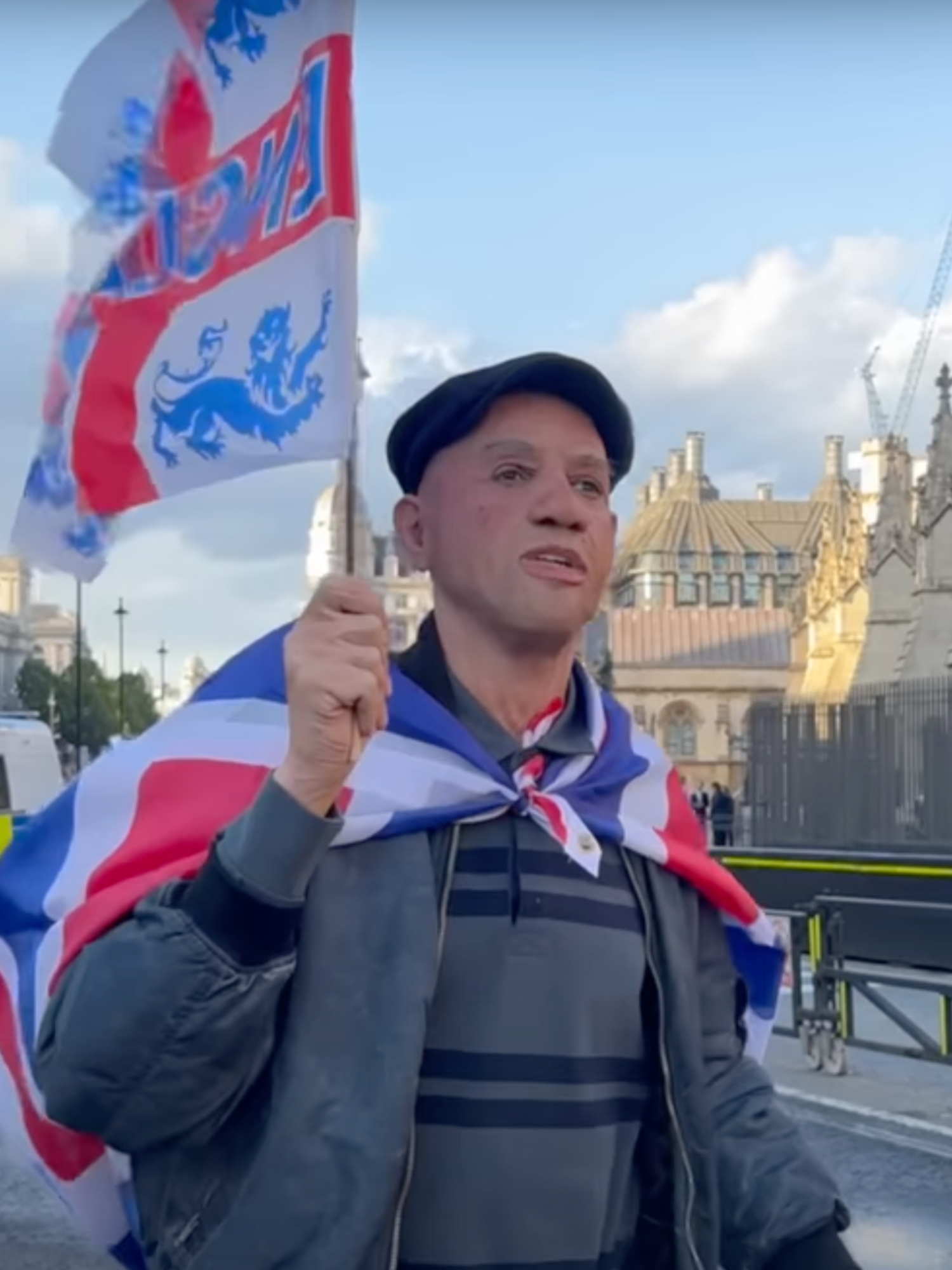 A man in a white, realistic face mask and flat cap carries an England flag down the road outside parliament, with two police vans in the background.
