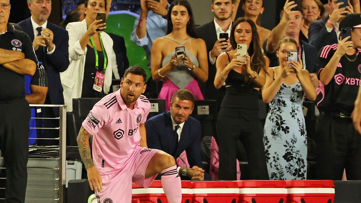 A man in pink football kit kneels on one knee, while fans film in the background