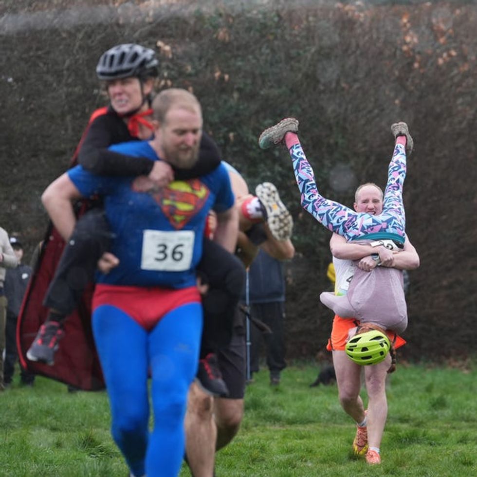 A man in the foreground wearing a Superman costume carrying his wife on his back while a man in the background carries his wife upside down in front of him