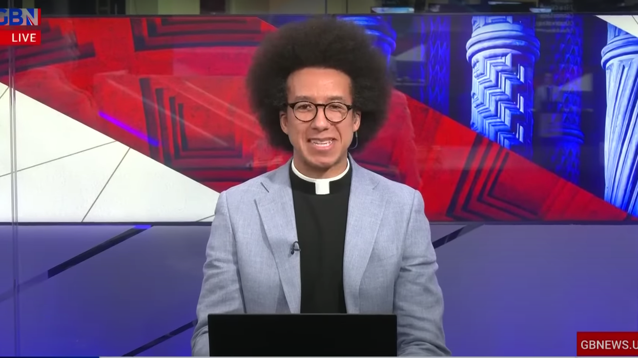 A man wearing glasses and a dog collar smiles on a TV news set