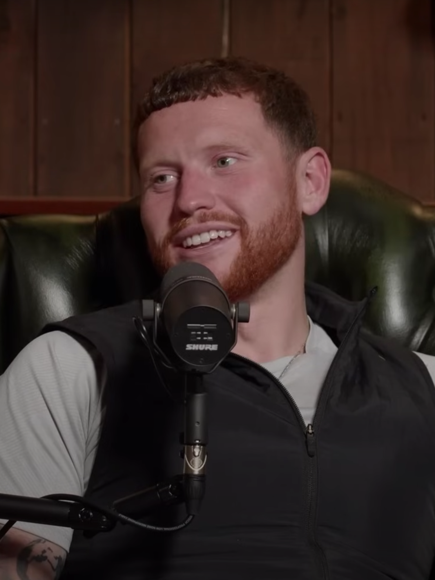 A man with ginger hair and a beard smiles in front of a microphone