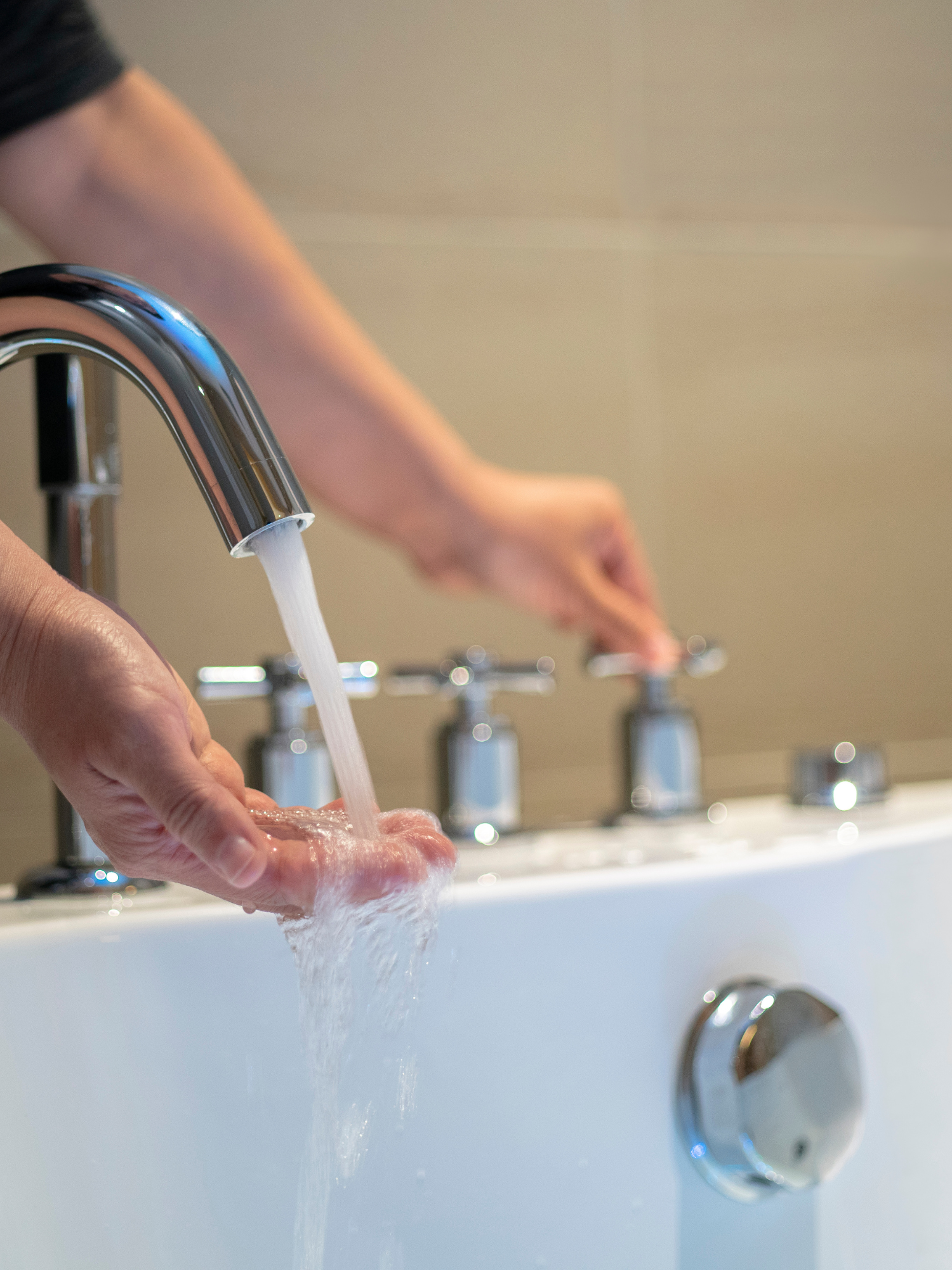 A pair of light-skinned hands over a white bathtub. The left adjusts the taps, while the right is testing the water temperature.