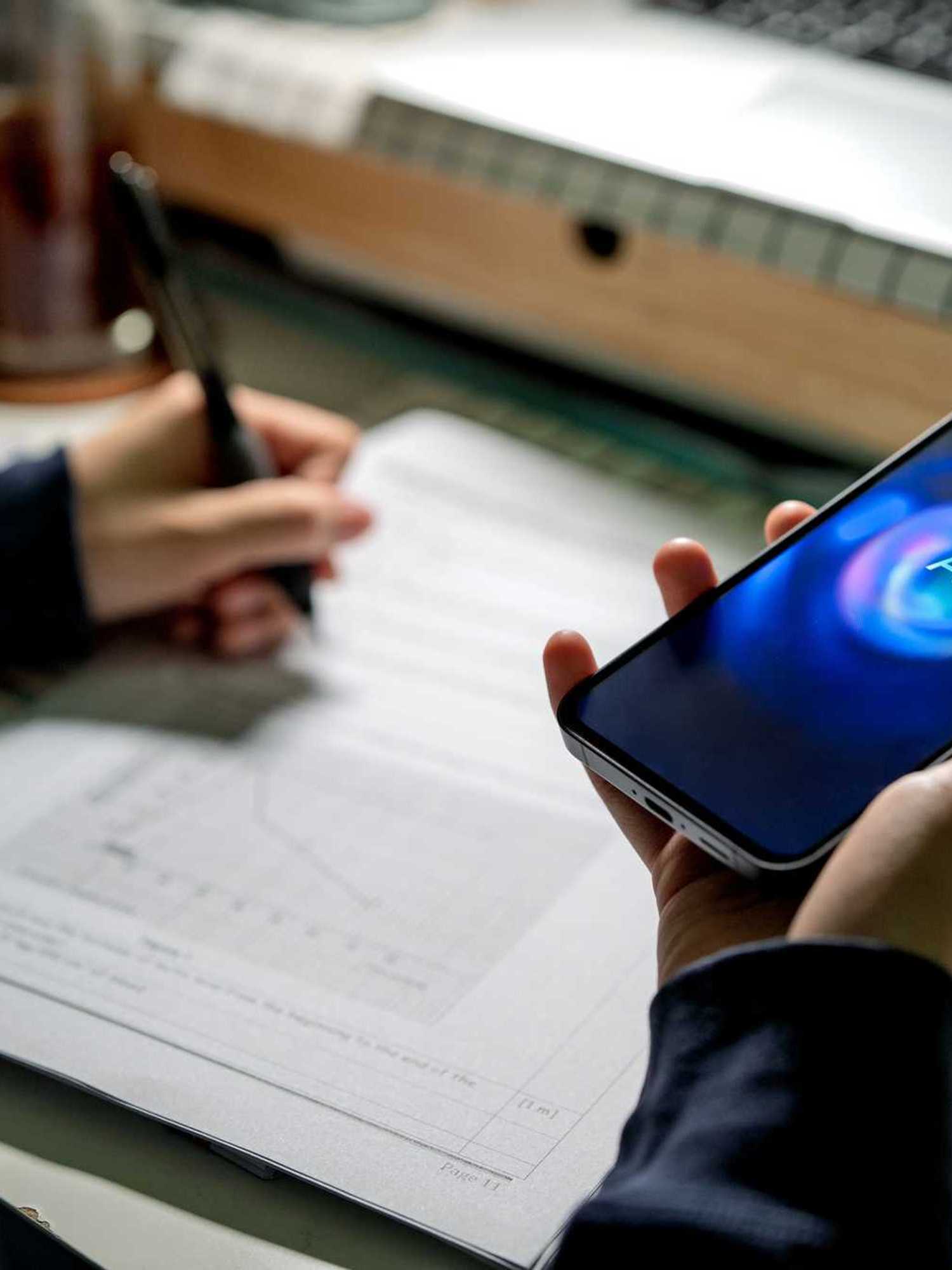 A person holds a phone displaying an AI chatbot in their right hand, while taking notes at a desk and writing with their left hand.