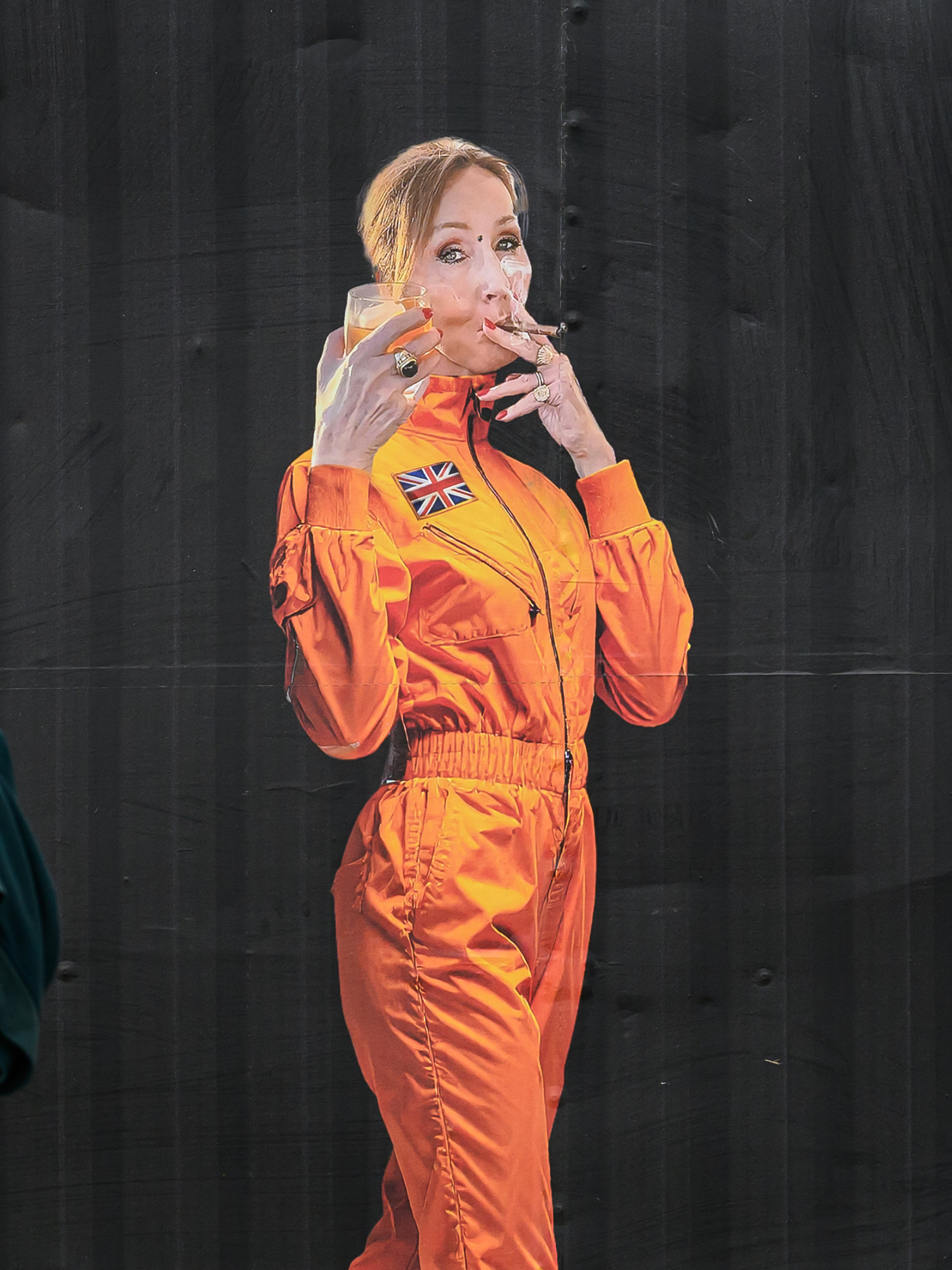 A poster at Glastonbury of JK Rowling in an orange space suit, holding a glass in her right hand and smoking a cigarette in her left hand.