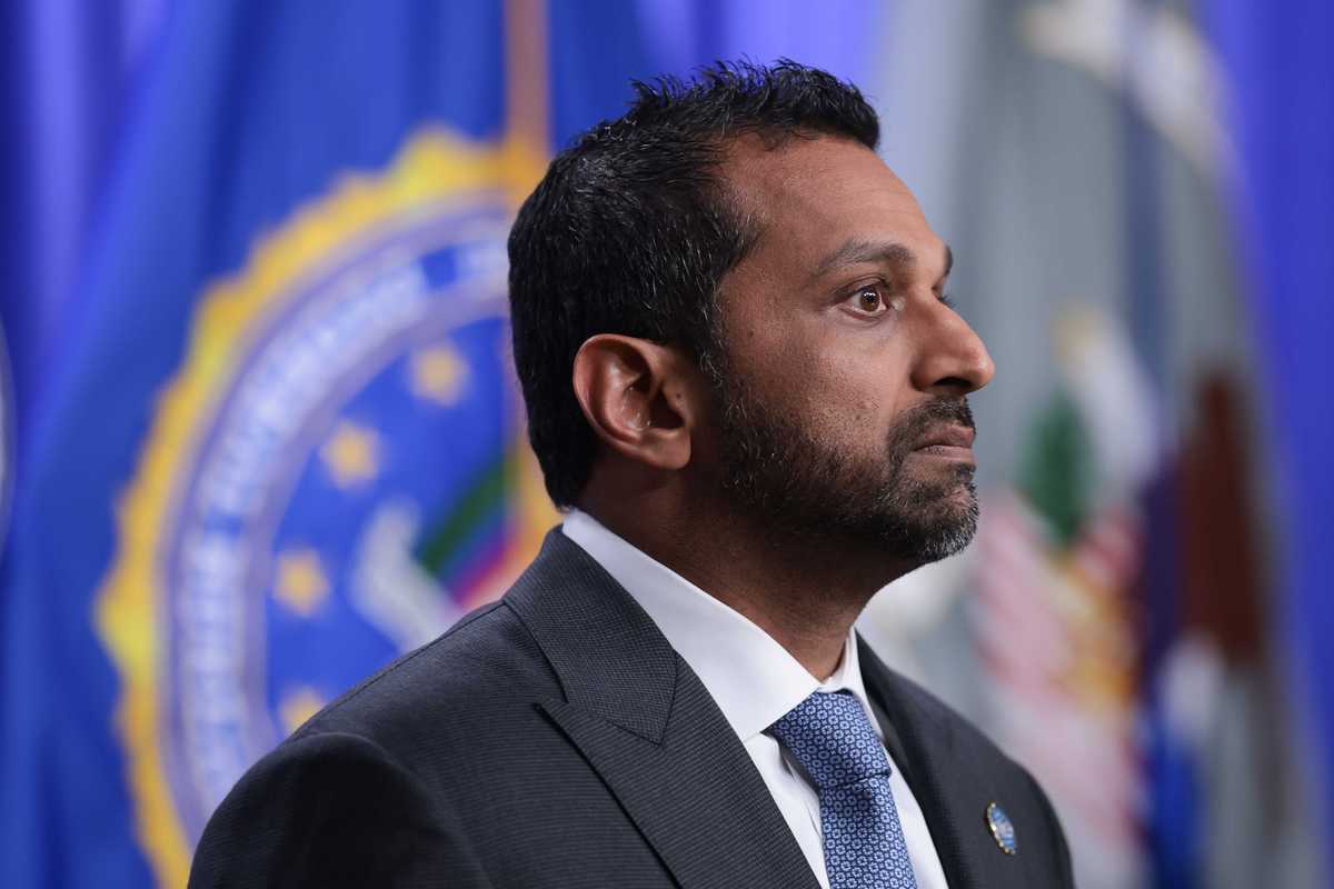 A side-on photo of Kash Patel, a bearded brown man with short black hair and a black suit.