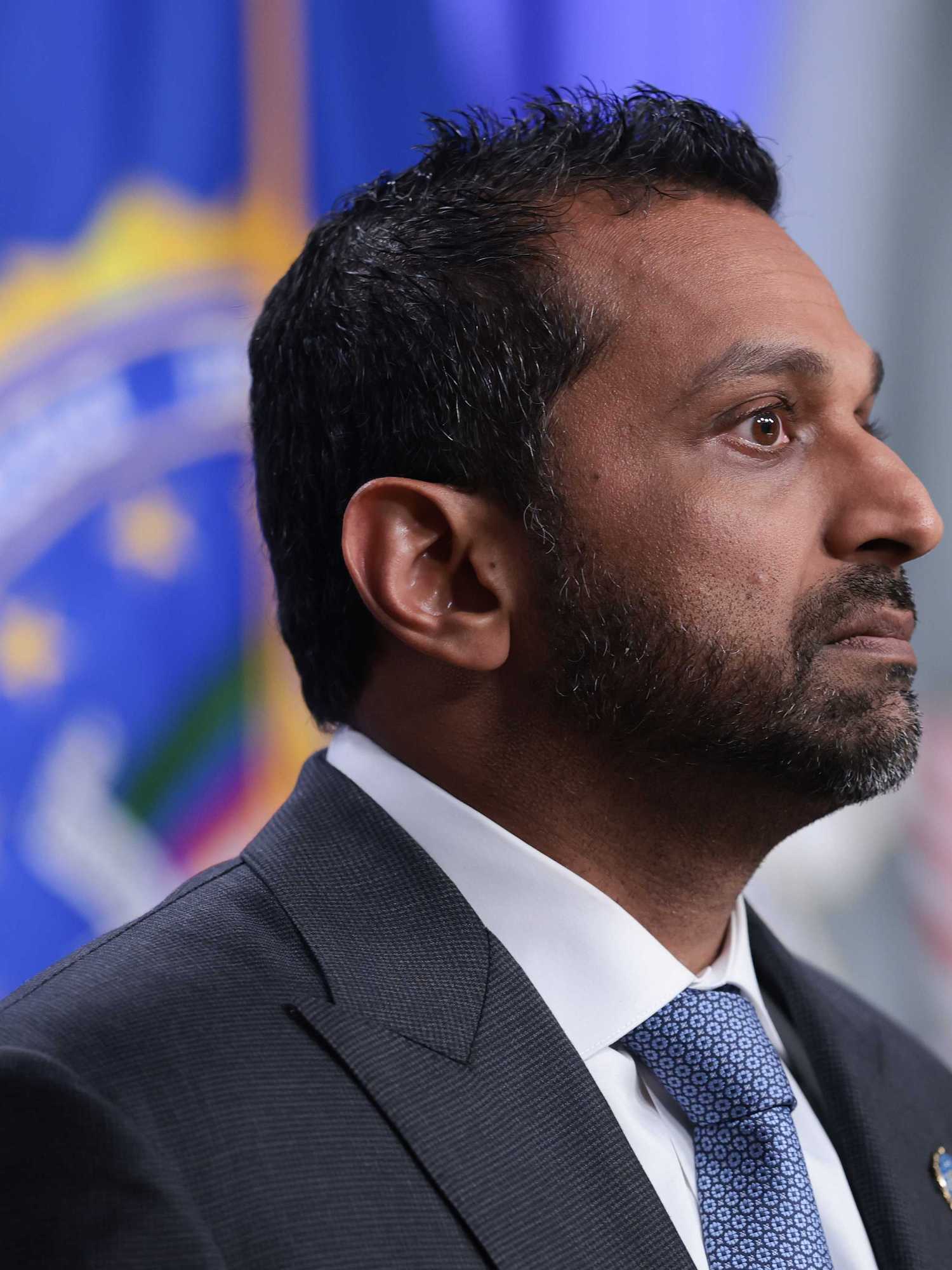A side-on photo of Kash Patel, a bearded brown man with short black hair and a black suit.