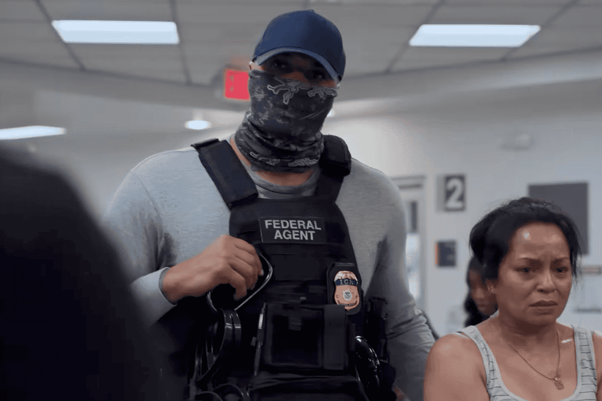 A still from The Pitt showing a male ICE agent, in a blue cap with a grey scarf covering his mouth and a bulletproof vest, standing next to a brown woman with a white vest and short black hair.