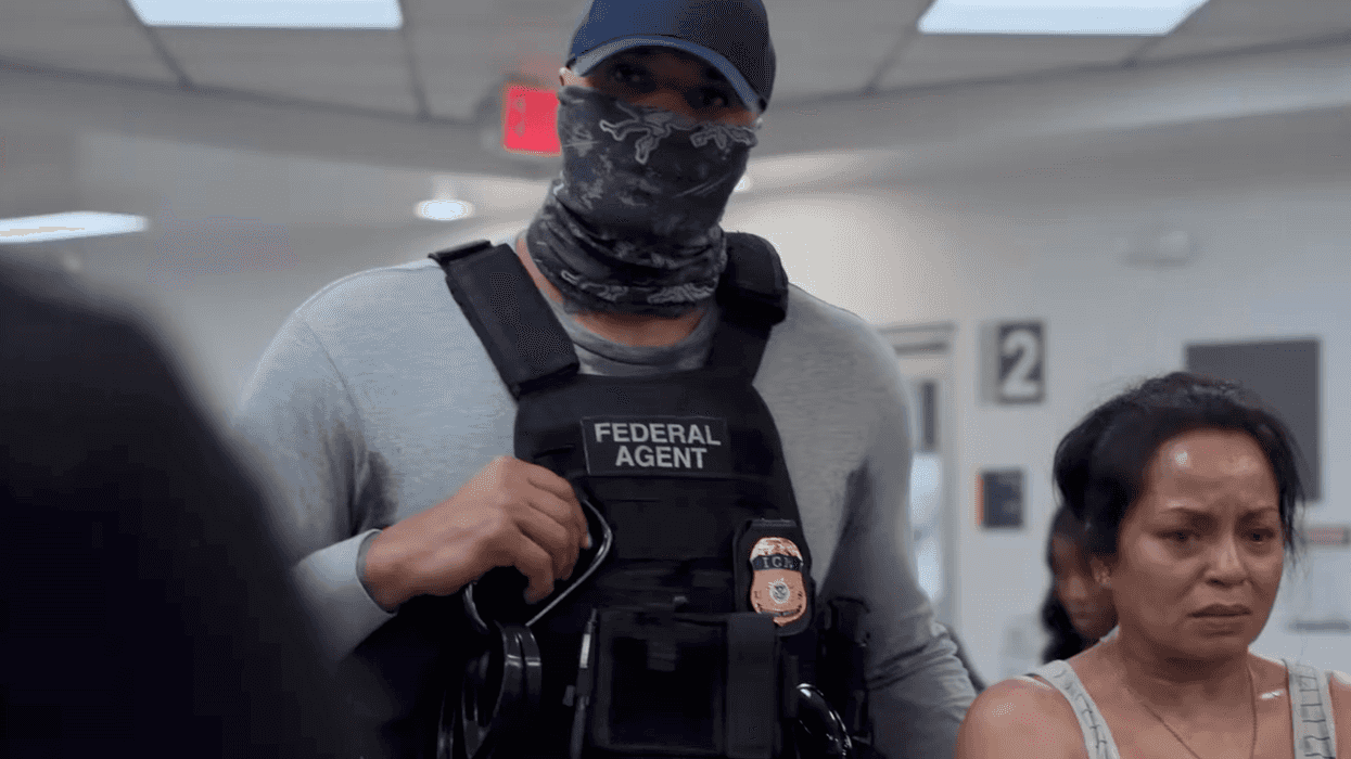A still from The Pitt showing a male ICE agent, in a blue cap with a grey scarf covering his mouth and a bulletproof vest, standing next to a brown woman with a white vest and short black hair.