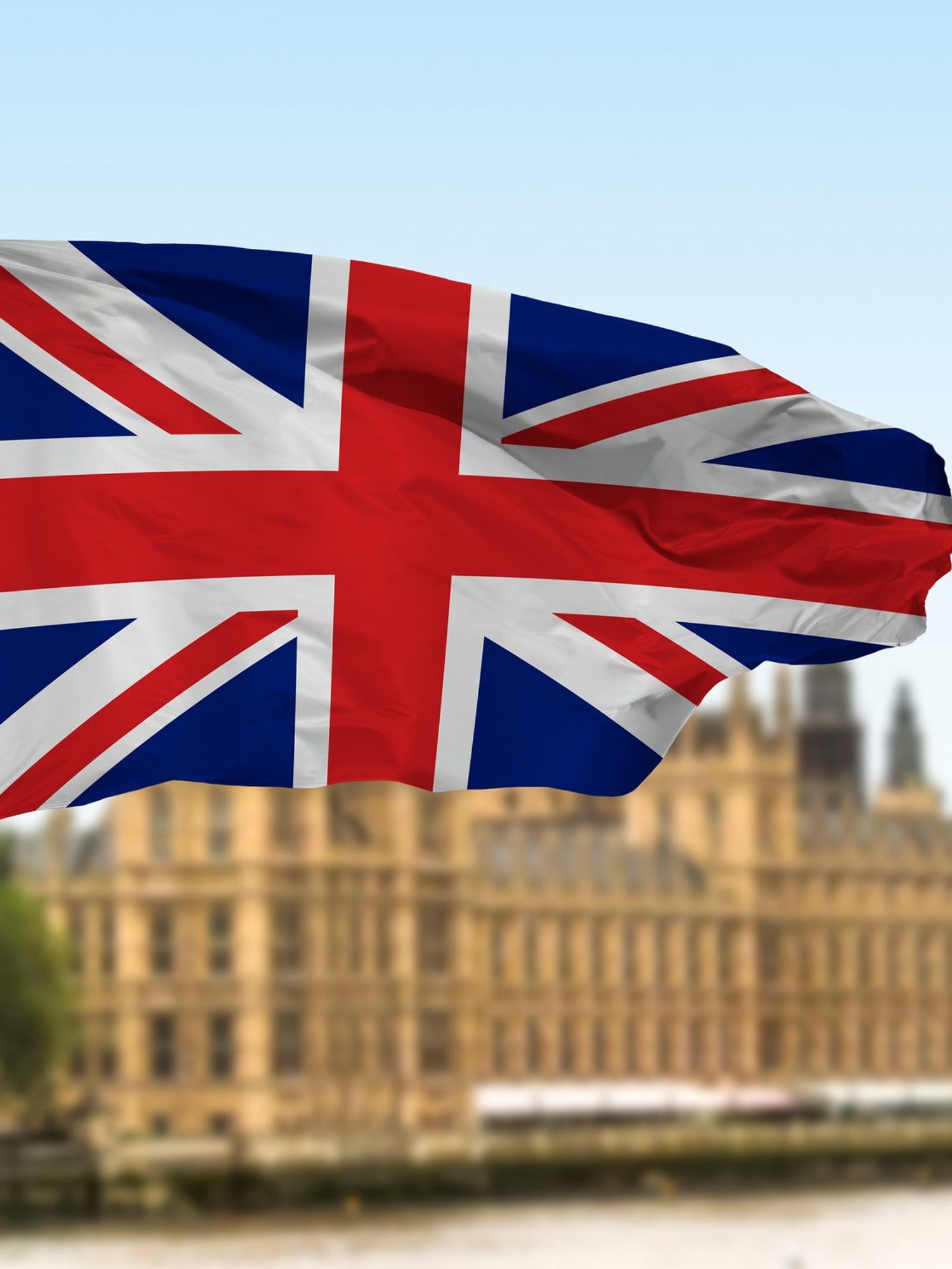 A Union Jack flag on a white pole waving in the wind, with the Houses of Parliament out of focus in the background behind it.