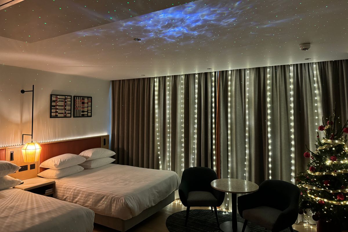 A view of a festive suite at the hotel. There are fairy lights on the curtains and a Christmas tree tucked in the corner away from the bed.
