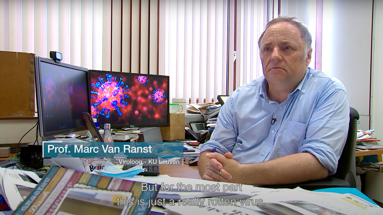A white man in a light blue collared shirt sits in front of a desk with sheets of paper scattered over it. He has two PCs in front of him and his hands are clasped together in front of him.
