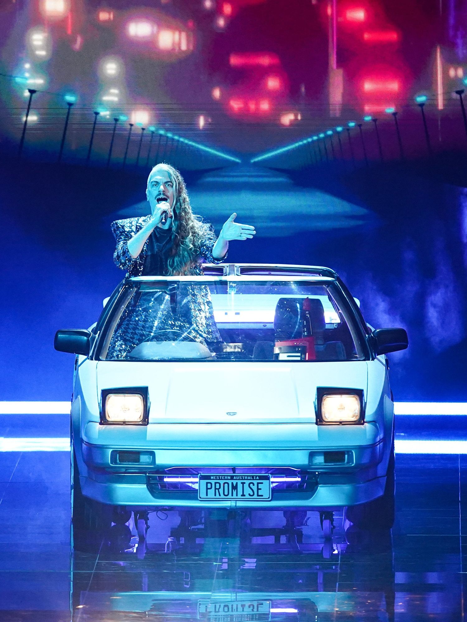 A white man with long blonde hair sings in the driver's seat of a stationary car on stage at the Eurovision Song Contest.