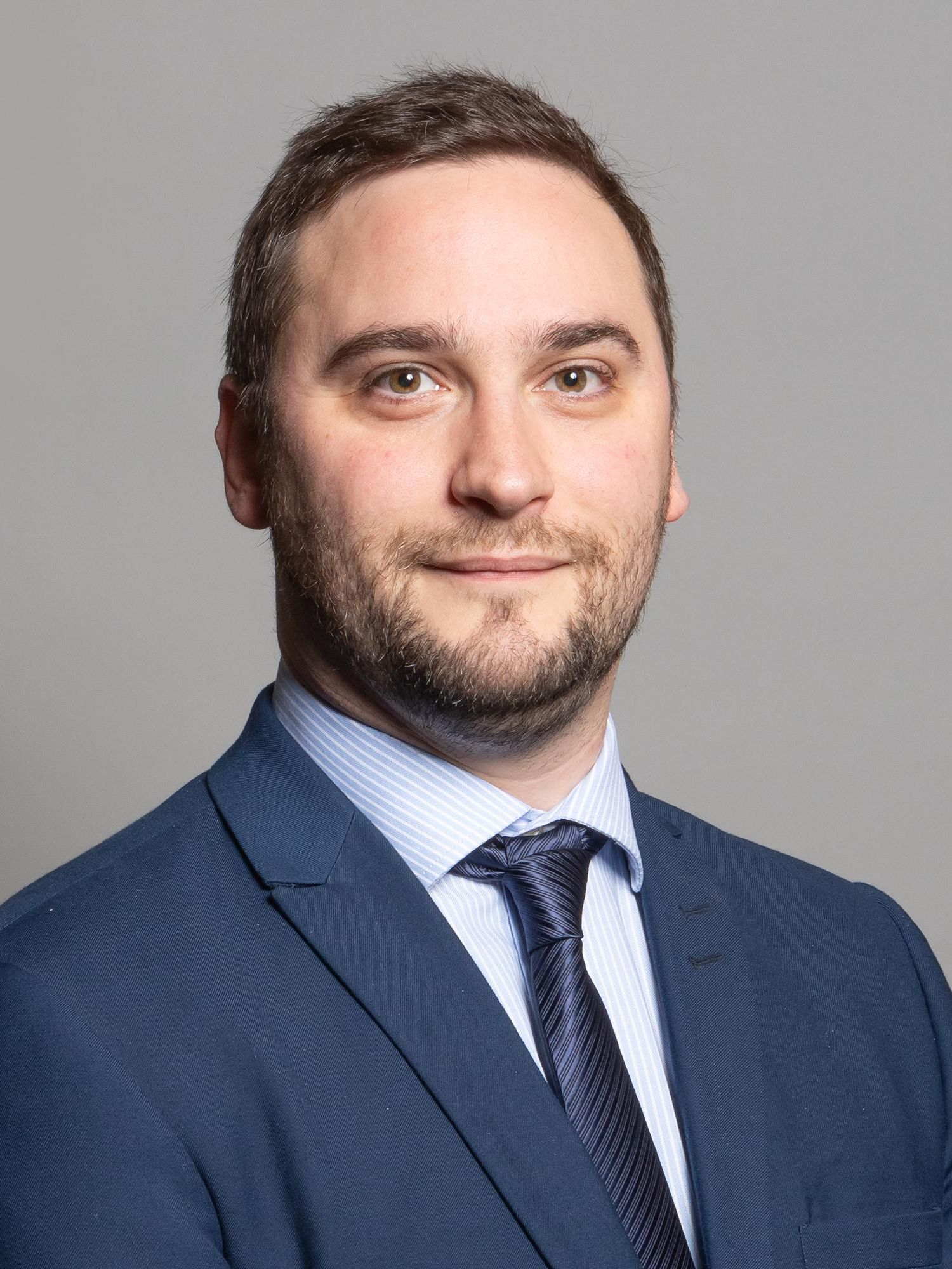 A white man with short black hair and a beard smiles in an official portrait. He wears a blue suit and tie.