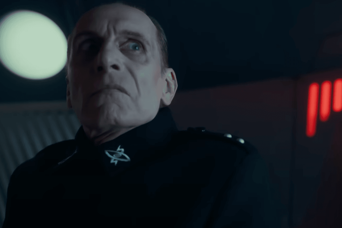 A white man with short grey hair and a black uniform gives a stern glare at someone off-camera.