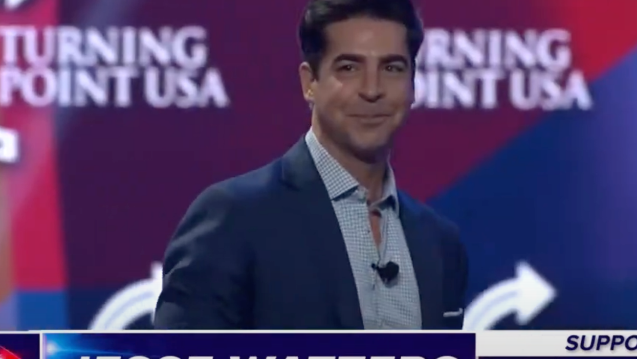 A white man with short, puffy black hair and a navy suit smiles on stage. Strapline text names him as Jesse Watters, and a screen behind him shows he’s talking at a Turning Point USA event.