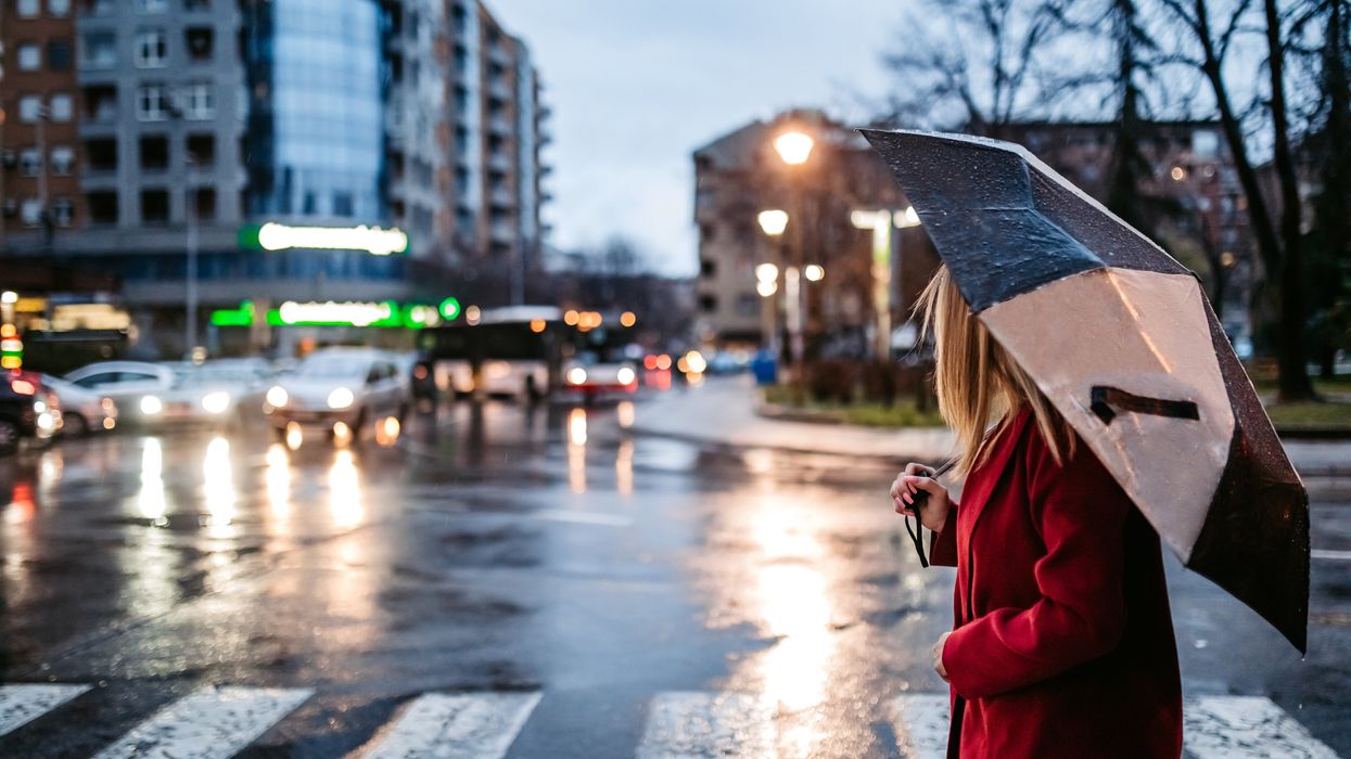 A white woman in a red coat and holding a black and white umbrella crosses a pedestrian crossing on a damp road.