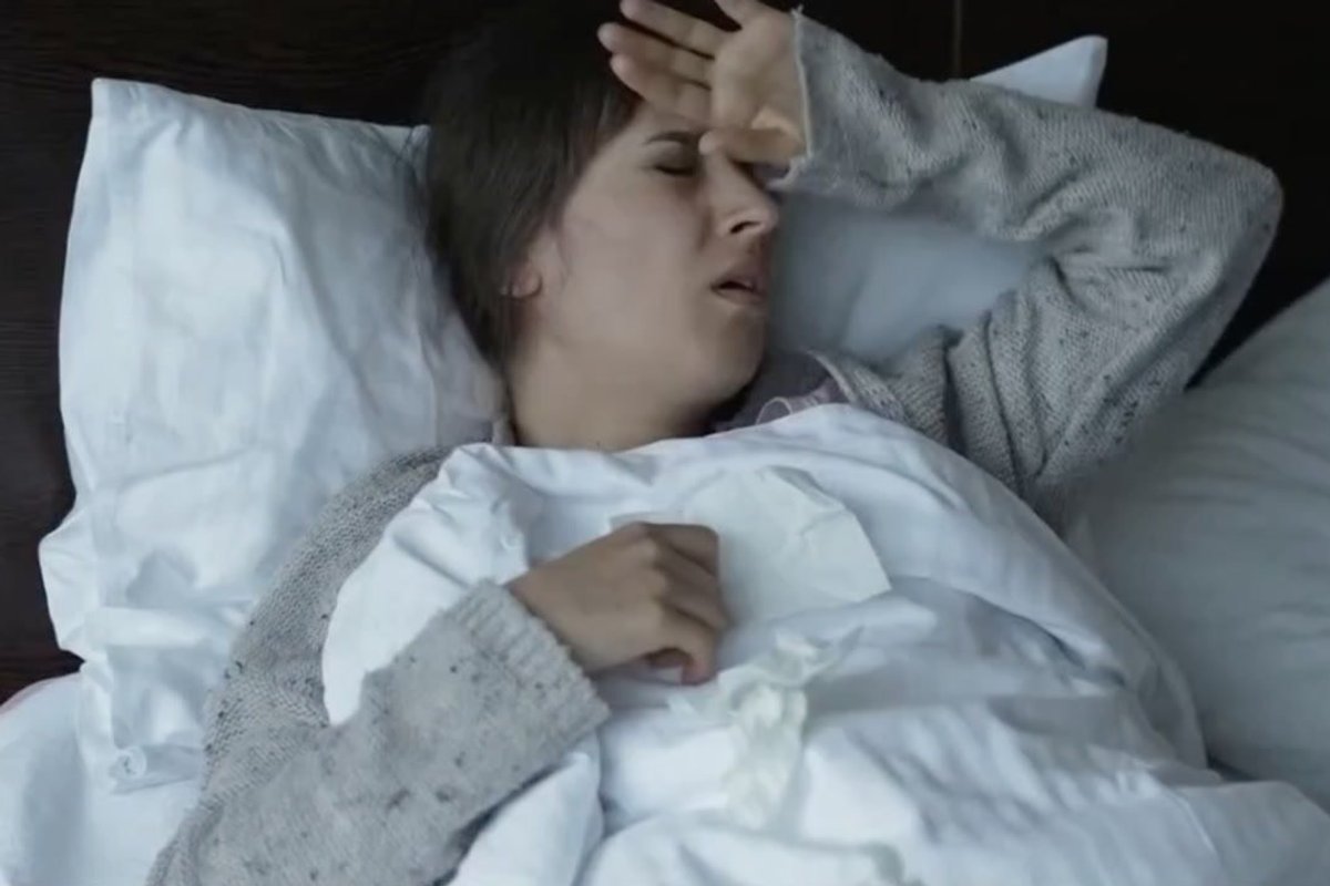 A white woman places her hand to her head as she sits in bed. She feels unwell.