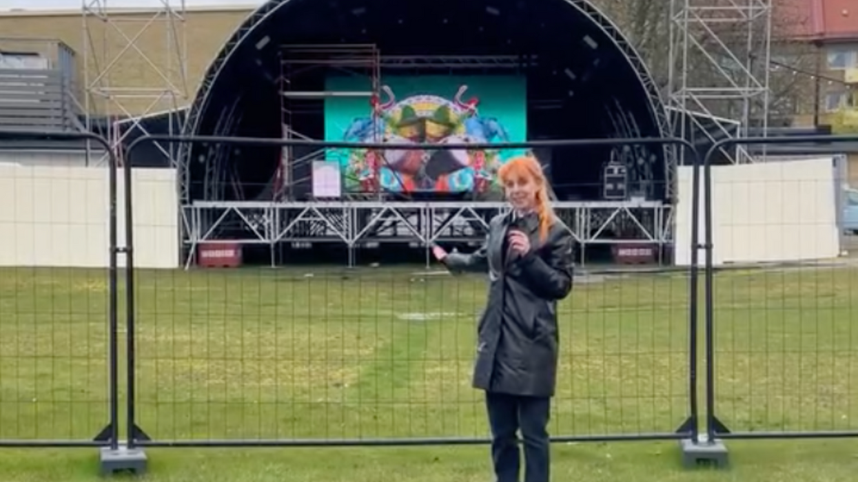 A white woman with orange hair gestures to a small stage with a curved roof behind gates behind her.