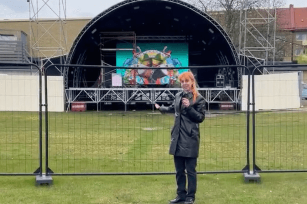A white woman with orange hair gestures to a small stage with a curved roof behind gates behind her.