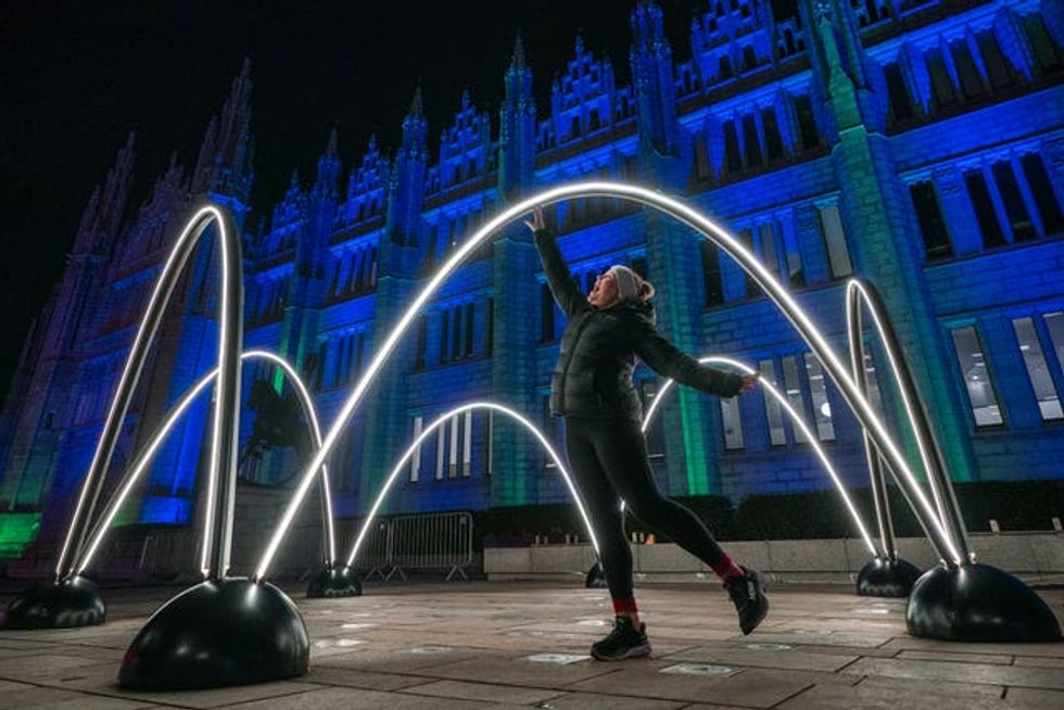 A woman reaches up to touch an arc of light, in front of a building lit up in blue