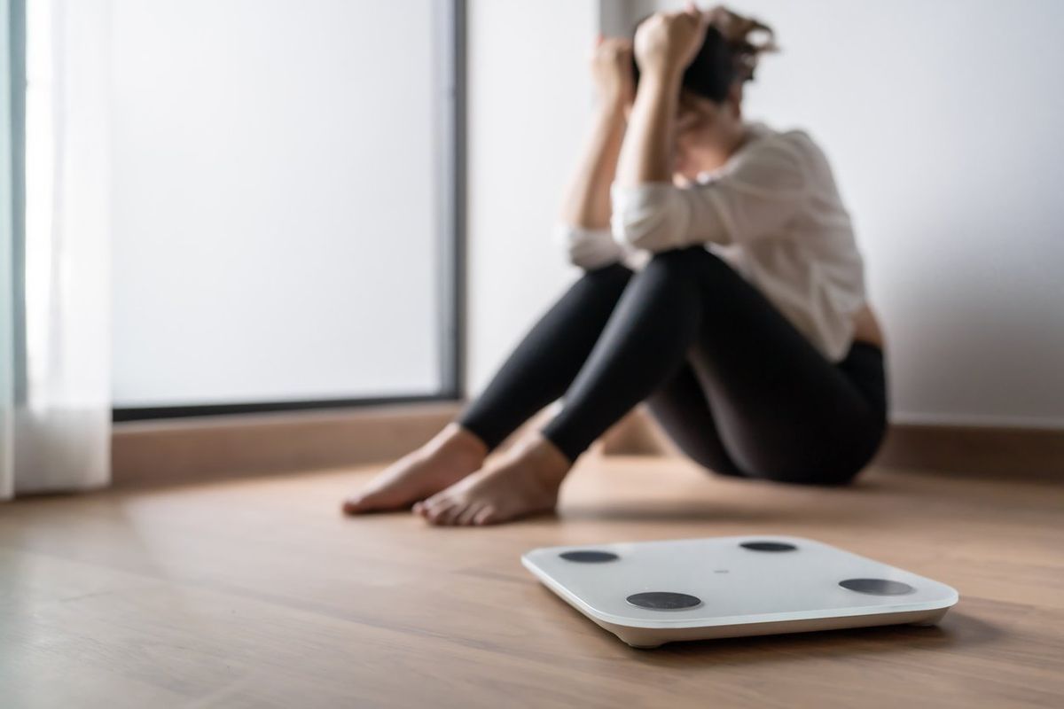 A woman sits looking distressed on the ground beside a set of bathroom scales