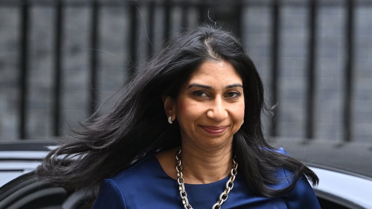 A woman with dark hair and a thick chain necklace smirks from in front of a car