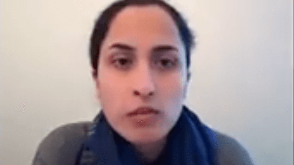 A woman with short curly hair is on a Zoom call. She has a blue scarf around her neck and wears a grey jumper. She is against a grey background.