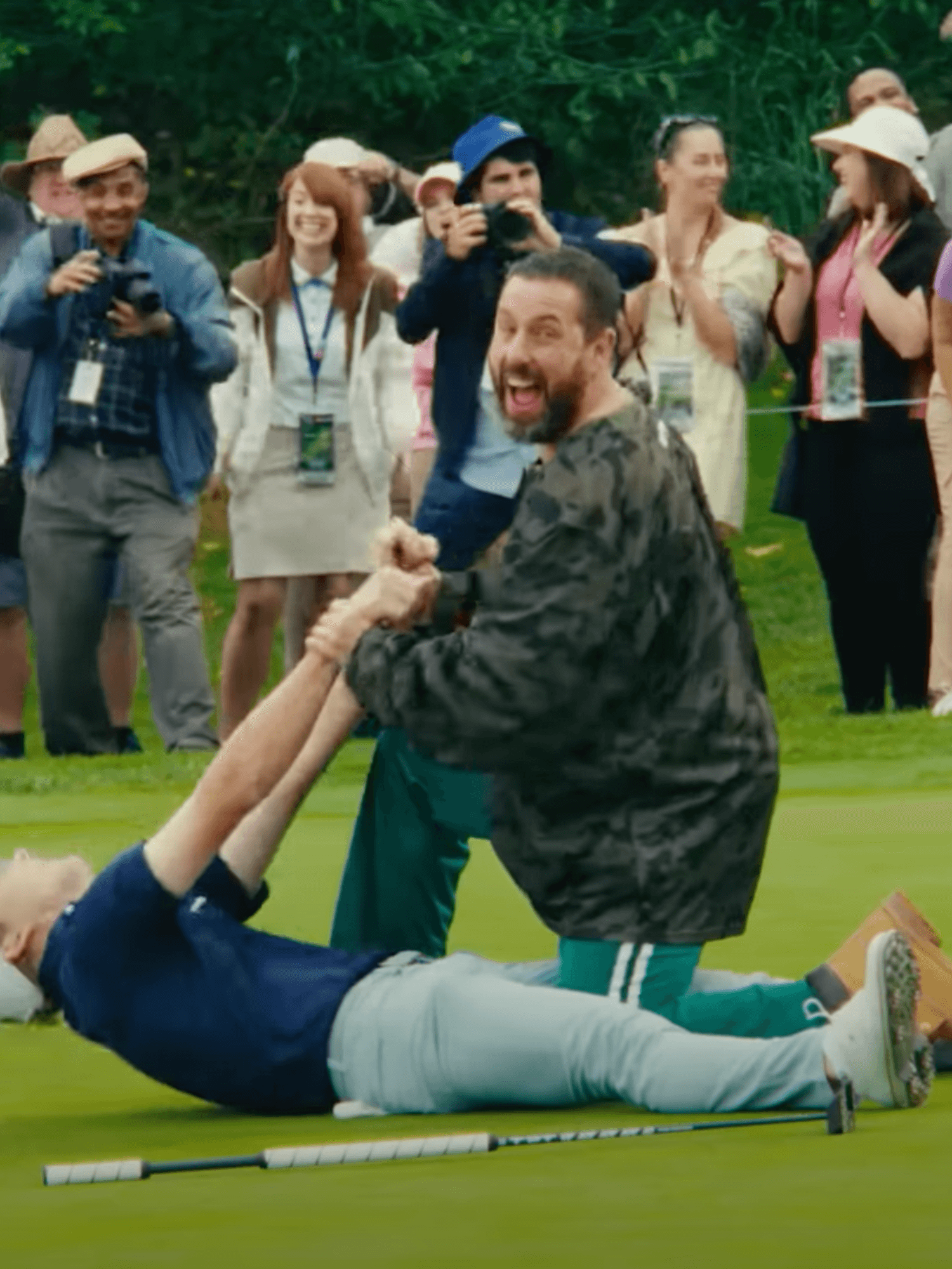 Adam Sandler as Happy Gilmore, smiling as he pulls the arms of another golfer lying down on a golf course.