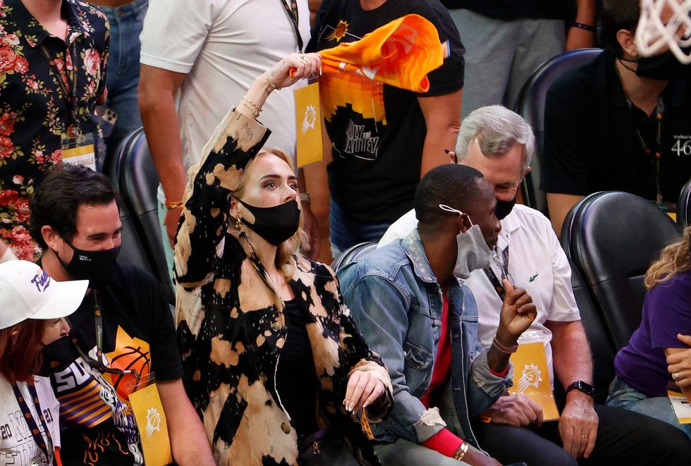 Adele is sat wearing a black and light orange jacket, waving an orange towel in the air with her right hand. She\u2019s watching a basketball game.
