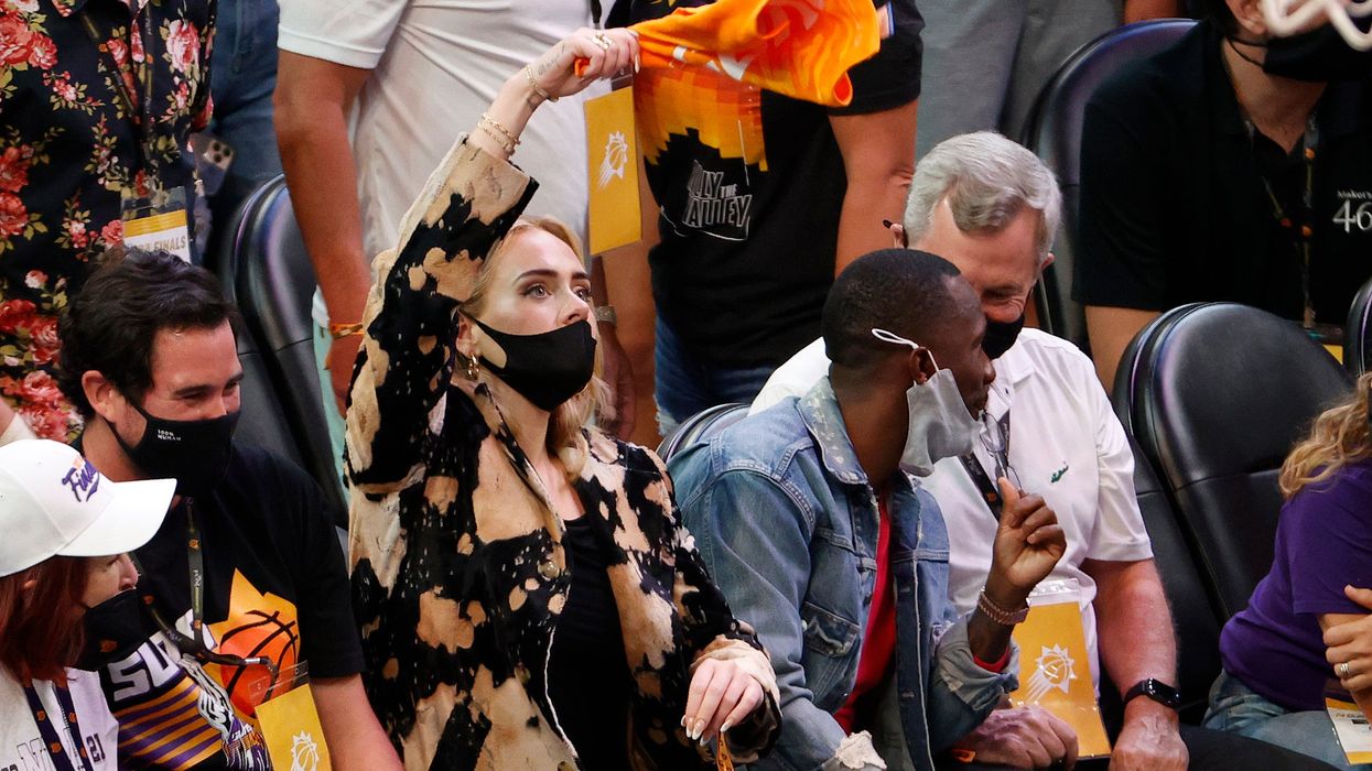 Adele is sat wearing a black and light orange jacket, waving an orange towel in the air with her right hand. She’s watching a basketball game.