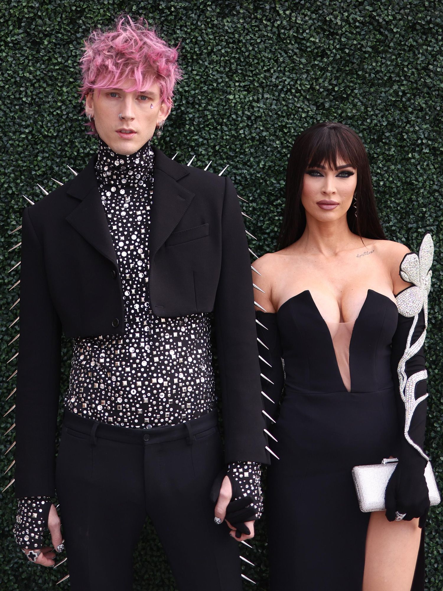 Against a green bush background stand Machine Gun Kelly, a white man with pink hair and a spiky black outfit, and Megan Fox, a white woman with long brown hair and a black dress. They have neutral expressions on their face.