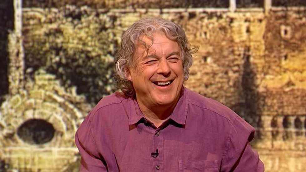 Alan Davies laughing during an episode of QI. He's a white man with short silver hair and wearing a purple collared shirt.