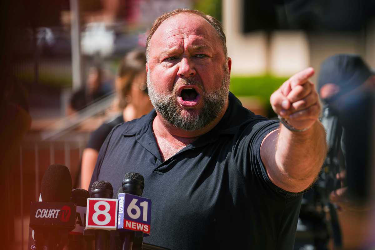 Alex Jones, a bearded white man with grey hair and a black shirt, points and shouts angrily at someone off-camera to his left.